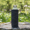 Szklana butelka wielorazowa w ciemnoszarym, filcowym pokrowcu z jasnym paskiem i uchwytem ze skóry, stojąca na kamieniu na tle zielonego lasu.
