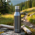 Szara, matowa butelka termiczna Forever ze stali nierdzewnej, stojąca na mchu i drewnie w plenerze. Solidny, minimalistyczny design ze srebrnym korkiem.