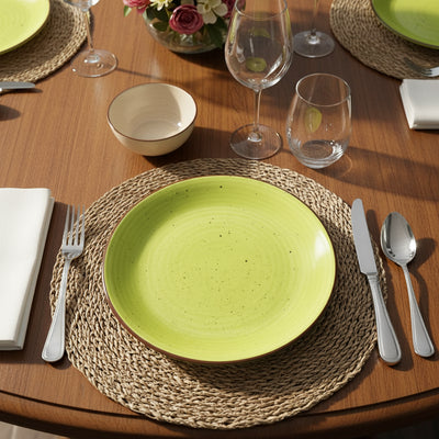 Okrągły, jasnozielony talerz deserowy Tognana z brązowym rantem i ciemnymi nakrapianiami. Leży na plecionej podkładce na drewnianym stole w otoczeniu sztućców i szkła. Idealny do rustykalnych aranżacji.